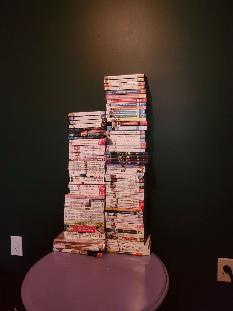 two tall piles of manga on a small round purple table, against a dark green wall. two white plated electrical outlets are visible on either side of the wall 