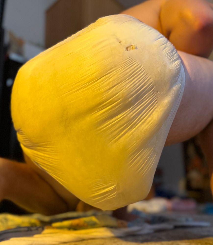 A boy with a massively full diaper is squatting in front of the camera. The diaper is incredibly used and noticeably discolored, to the point where you can see stains from the inside. The back of the diaper has a rip near the top from the weight of the destroyed diaper.