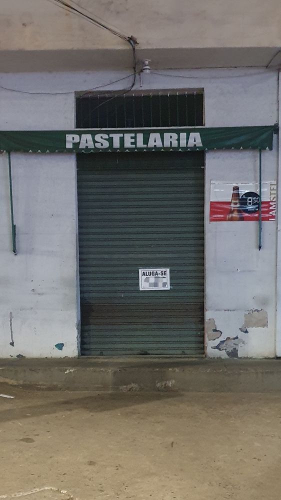 Fachada branca com uma porta de bar verde escura e um toldo verde escuro recolhido, onde se lê PASTELARIA. Partes da parede têm manxhad e a tinta descascando e há um anúncio de cerveja colado no lado direito