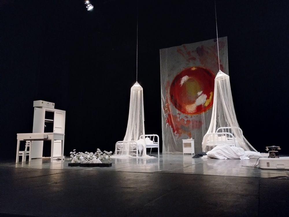 Escenario teatral al final de la obra Ícaro en el Teatro de la Ciudad, CDMX. Solo muebles blancos: un armario, un escritorio, una silla pequeña, dos camas de metal cubiertas por mosquiteros. Entre ambas camas, una pintura gigante de un sol, radiante y cálido. Una maleta en una esquina.