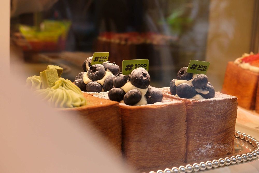 Close-up photo of a tray matcha cube croissants and blueberry cube croissants in a bakery display window. A small paper label stuck to the top of each croissant reads, "#1000 Bread"