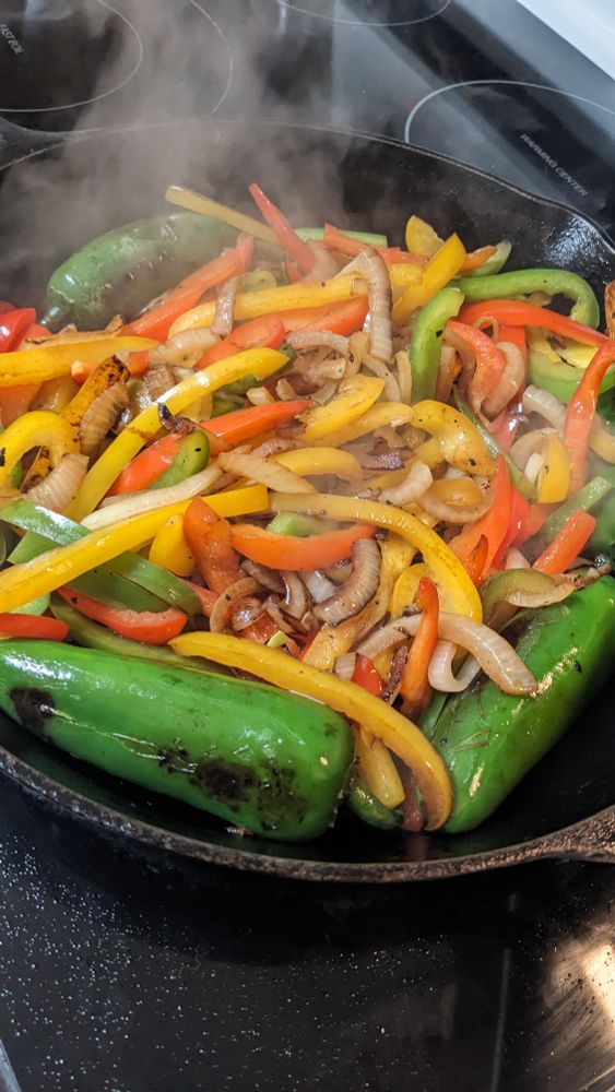 Red, yellow and green bell peppers, onions and jalapenos brown in the cast iron. Steam rises from the vegetables. 