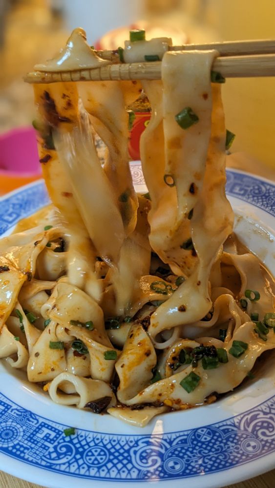 Chopsticks lifting long, hand-cut noodles in a sesame peanut sauce with chili crisp and scallions. Fat noodles reside in a blue and white Calamity Ware bowl. 