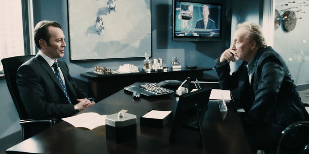 Screenshot from the movie Saw VI, featuring the character John Kramer aka Jigsaw sitting across a desk from health insurance executive William Easton. In this scene, Kramer is denied coverage for his cancer treatment.