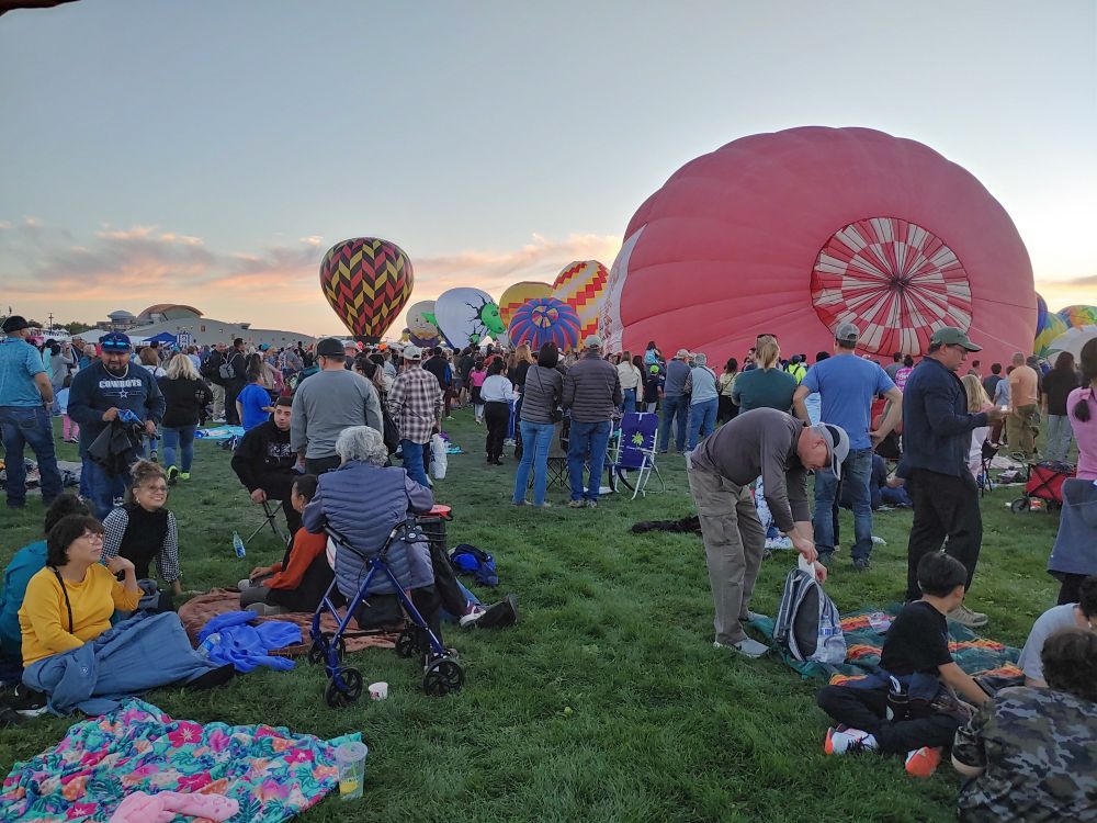 Hot air balloons being inflated in the back ground while a crowd of people watch from across a field in the foreground