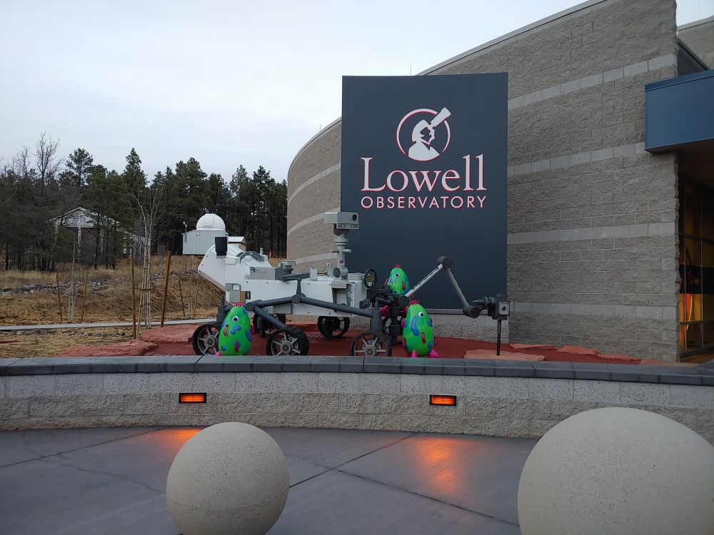 A 6-wheeled space-faring robot sitting in front of a grey sign with the works "Lowell Observatory" and a graphic of a man looking through a telescope. 
