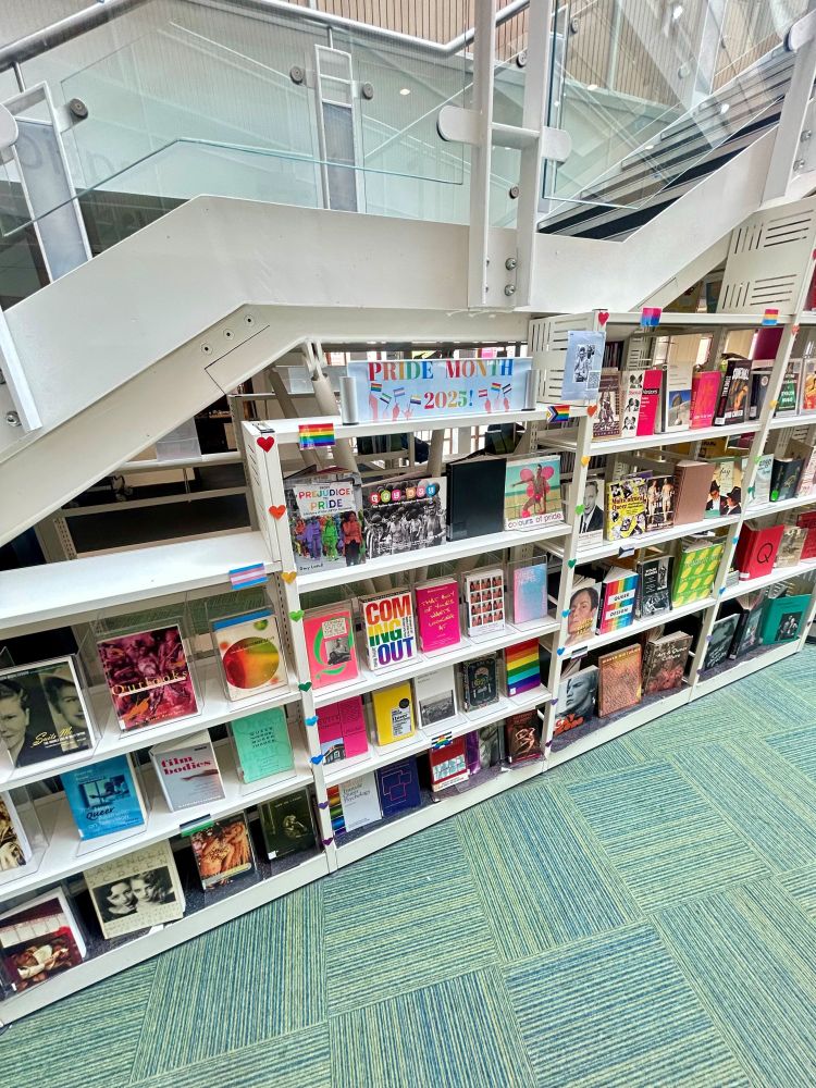 A book display in a library. A banner at the top reads "Pride Month 2025" in rainbow colours. There are rainbows and multi-coloured hearts across the book display. There are various texts on a shelf spanning the picture.