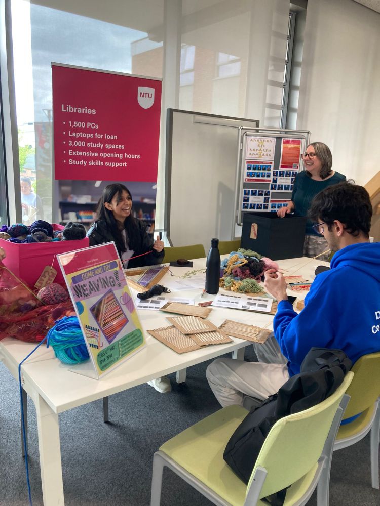 A group of people are seated around a table in a library, engaging in weaving. The table is covered with various materials including yarn, woven samples, and instructional sheets. A sign on the table reads "Come and Try Weaving." In the background, there is a large pink banner with the NTU logo that lists library services such as 1,500 PCs, laptops for loan, 3,000 study spaces, extensive opening hours, and study skills support.