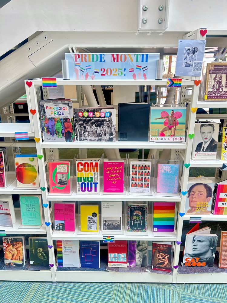 A book display in a library. A banner at the top reads "Pride Month 2025" in rainbow colours. There are rainbows and multi-coloured hearts across the book display. There are various texts on a shelf spanning the picture.