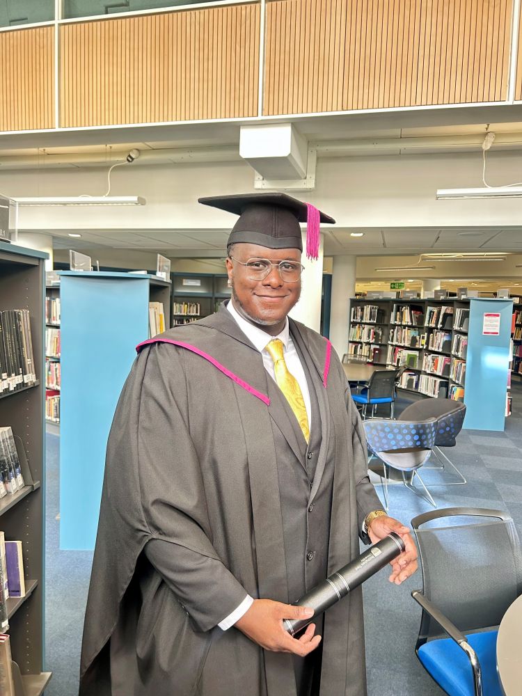 A person wearing a graduation cap and gown stands in a library. The individual is holding a rolled-up certificate in their right hand. The gown is dark with pink trim, and the person is also wearing a yellow tie. Bookshelves filled with books are visible in the background, along with some chairs and tables."