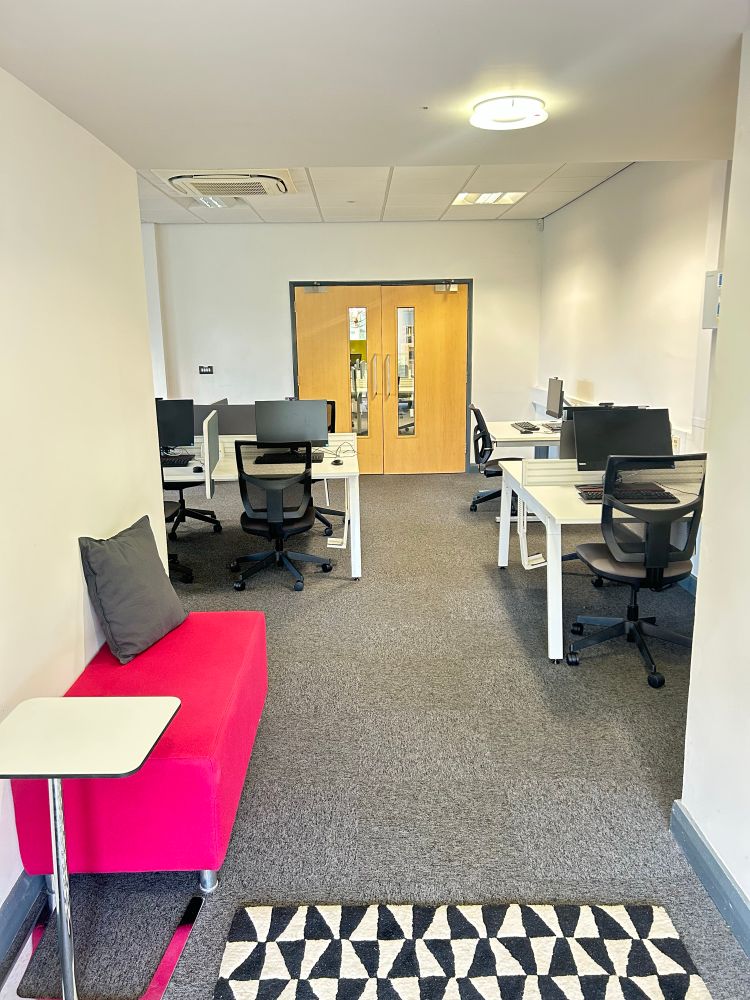 A modern office space with several workstations. Each workstation has a computer monitor, keyboard, and office chair. The room has white walls and a grey carpeted floor. There is a bright pink bench which a grey cushion on the left next to a small white table. At the far end of the room are double wooden doors with glass panels. 