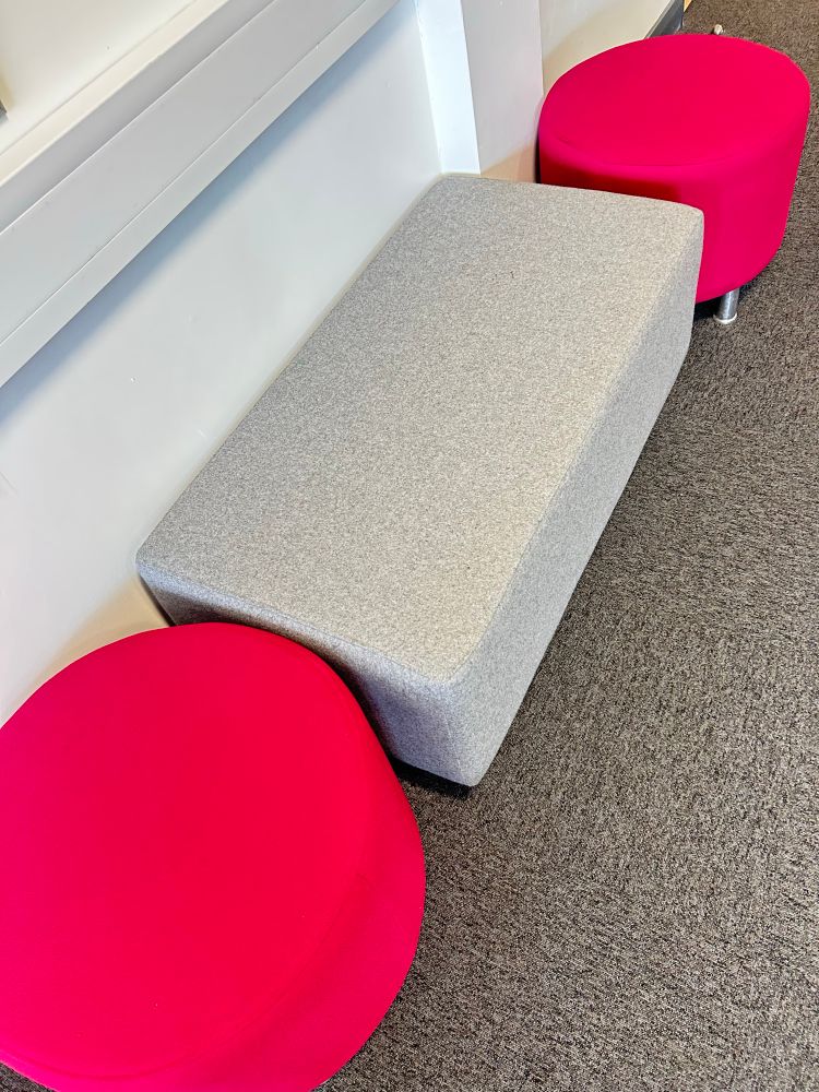A seating area with two round bright pink stools and one rectangular light grey bench. The furniture is set on a dark carpeted floor next to a white wall. 