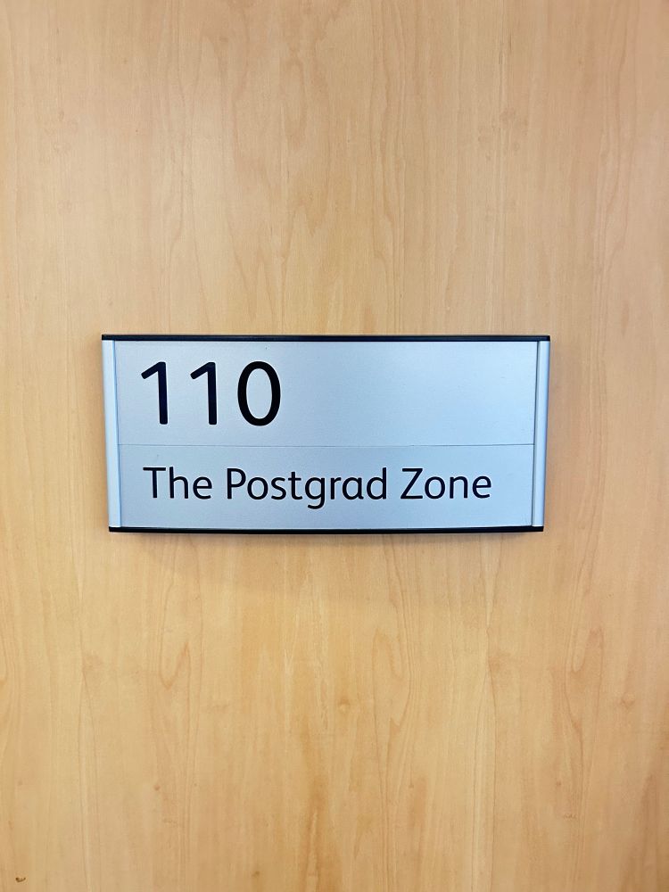 A wooden door with a silver and black sign that reads “110 The Postgrad Zone.”