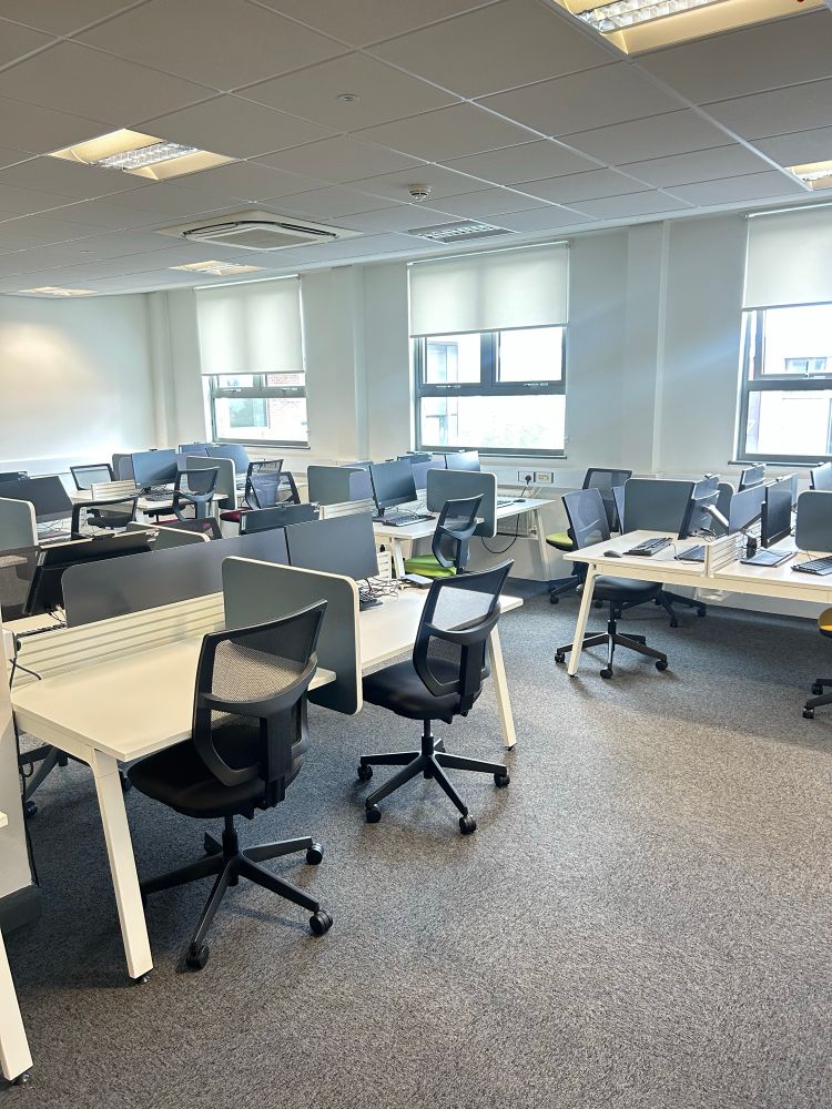 A modern office space with multiple workstations. Each desk has an office chair and computer monitors. The room is well-lit with natural light from the windows and ceiling lights. Desks are arranged in rows with partitions.