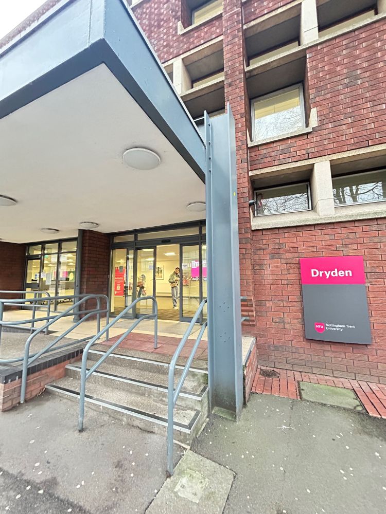The image shows the entrance to a building named "Dryden" at Nottingham Trent University. The entrance has a small set of stairs with metal railings leading up to glass doors. A pink and grey sign on the right side of the entrance reads "Dryden" with the Nottingham Trent University logo below it. The building is made of red brick, and there are several windows visible above the entrance.