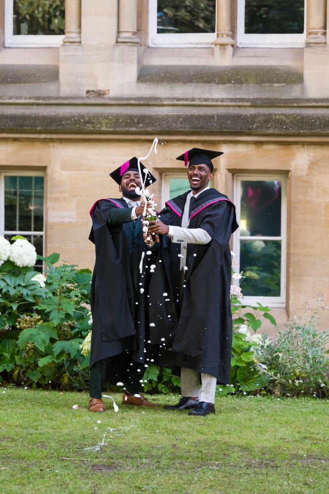 Two individuals in graduation gowns and caps stand on a grassy area in front of a building with large windows. They have wide joyful smiles with teeth showing. They are holding and spraying a drink, with the liquid visibly splashing out of the bottle. Plants and flowers are visible in the background near the building.