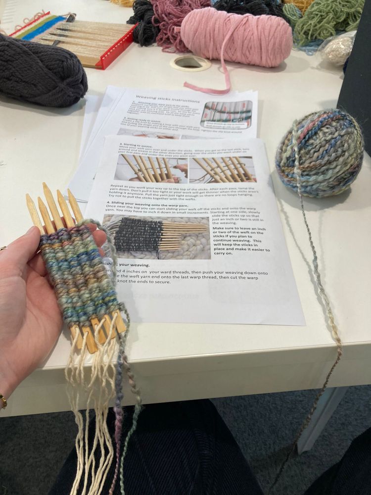 A person is holding a small weaving project on wooden sticks. Various balls of yarn (in many colours) and weaving tools are spread out on the table. The background includes instructional papers with text and images related to weaving.