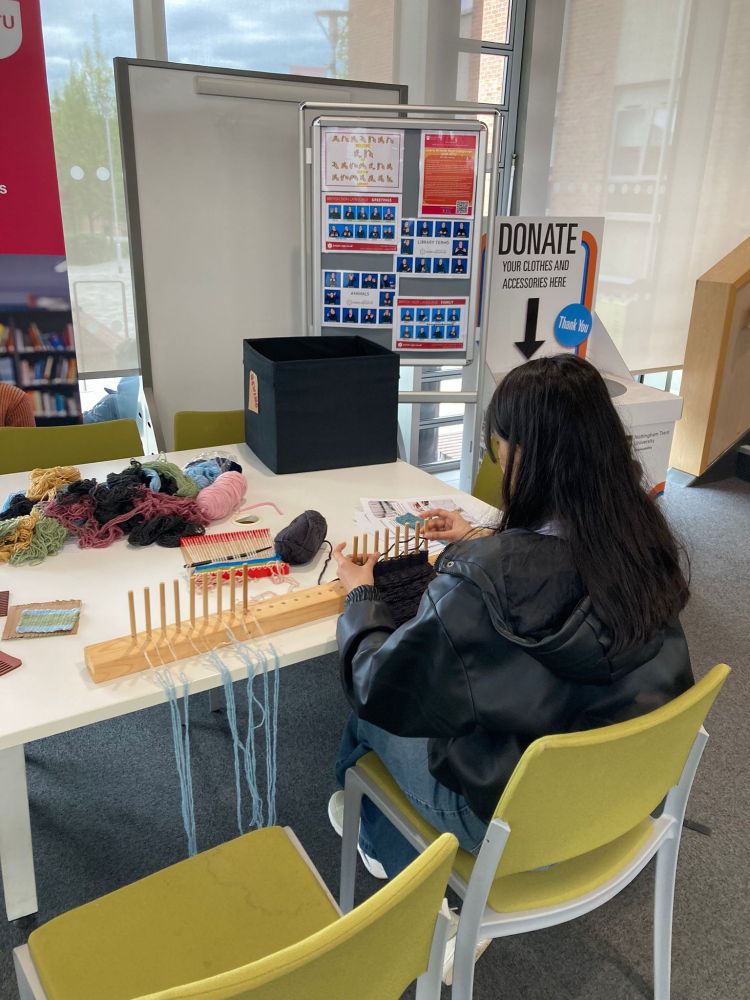 A person is seated at a table in the library, working on weaving using a wooden loom with multiple pegs. Various yarns and knitting materials are spread out on the table. In the background, there is a donation box and an informational board displaying images and text related to knitting or weaving techniques.