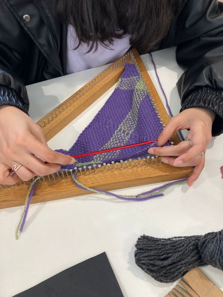 A person is weaving on a triangular wooden loom, using a red needle to weave purple and green yarn. The person's hands are visible, holding the needle and working with the yarn. There is also a bundle of dark gray yarn on the table next to the loom.