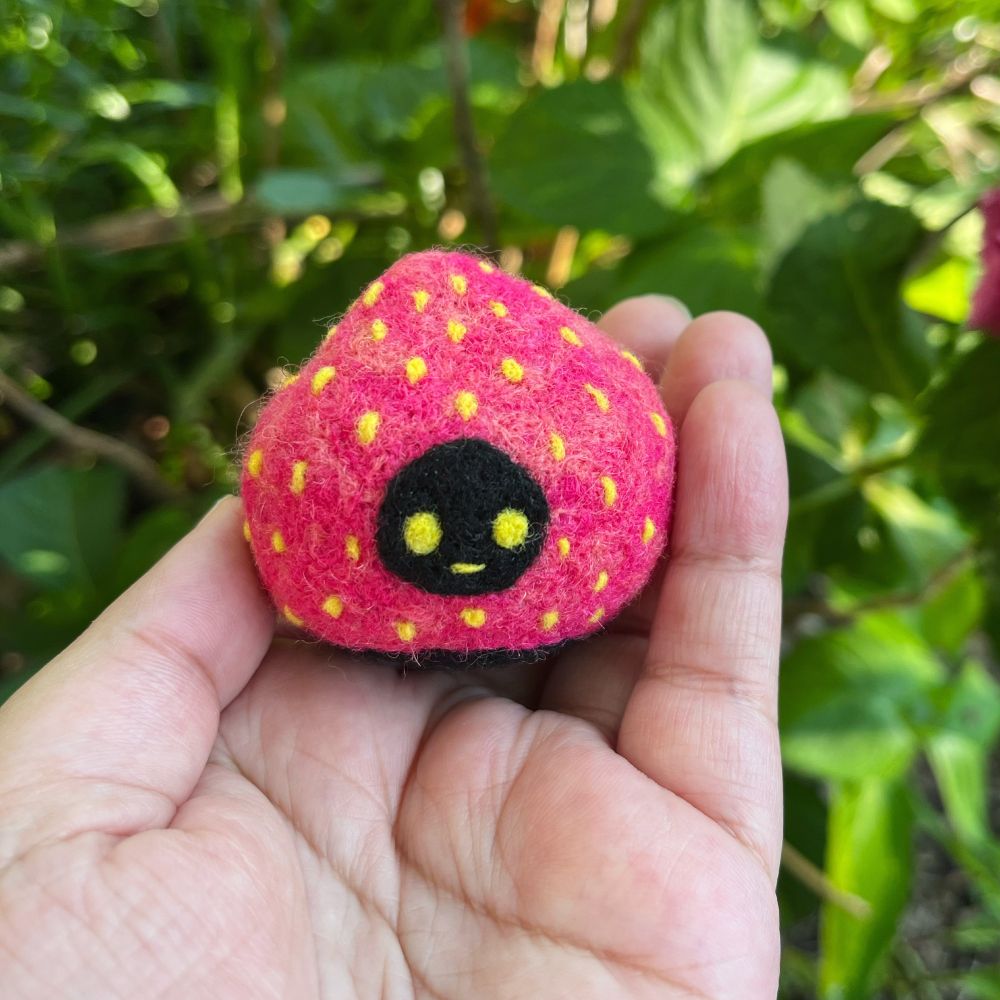 Photo of hand holding a small needle-felted sculpture of a creature living inside a strawberry. Her name is Candy