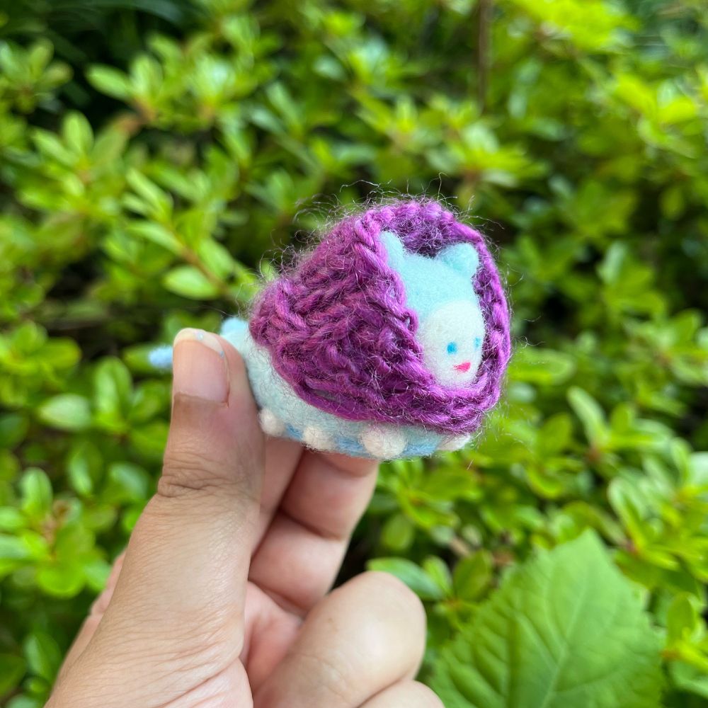 Photo of hand holding a small needle-felted sculpture of a light-blue caterpillar-like creature, who is wearing pink lipstick and a purple hand-knitted shawl. Her name is Beba