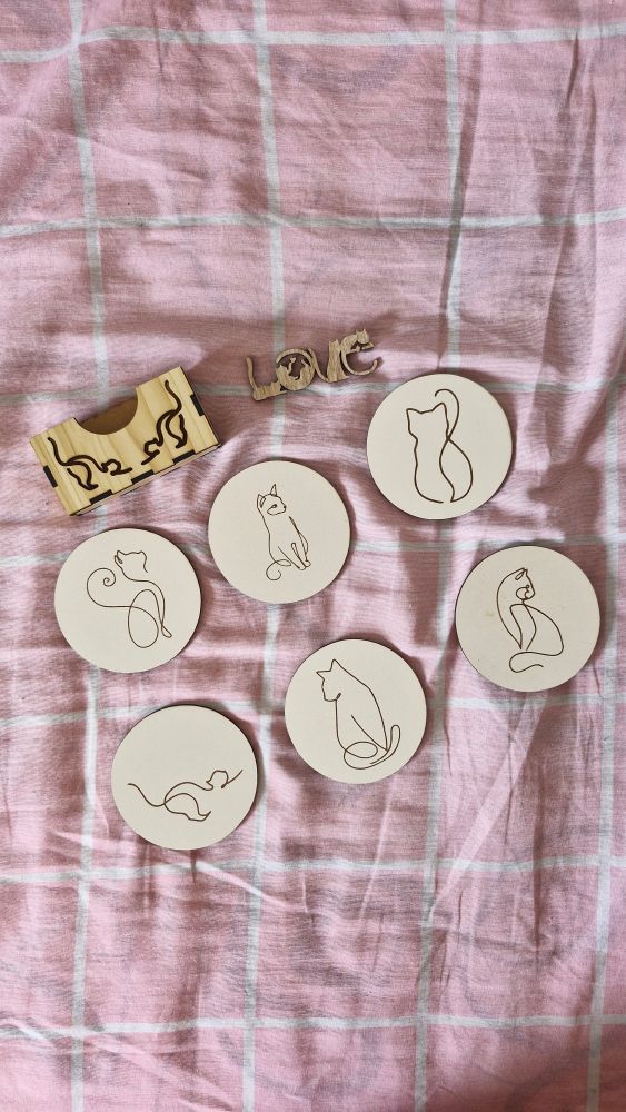 A mesma caixinha da foto anterior, com os discos, que são descansos de copos, para fora. Cada um tem um ten um desenho de silhueta de gato em uma posição diferente. Sentado de perfil, sentado de frente olhando para o lado, de costas, se espreguiçando, etc. Outro adereço de madeira tem a palavra "love" estilizada em forma de quatro gatos em diferentes posições felinas