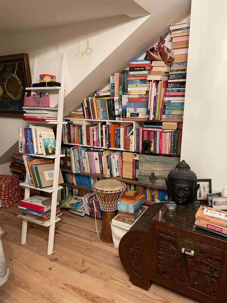 A pile of shelves, books and travel memories, including a buddha head in the foregroubd, a djembé drum, a pile of frames and 1001 night slippers. In front on the right a large woodcarved chest. On the left a leaning slant bookcase with mainly childrens books. 