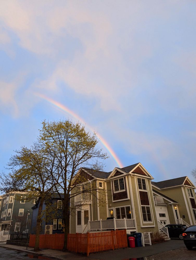 Houses and trees with rainbow 🌈 in the blue sky. There is a faint second rainbow arc above. 