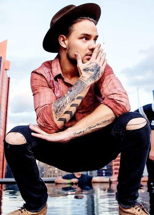 Liam Payne showing off his leg strength in 1D days wearing a hat, black skinnies, brown shoes and a plaid shirt 