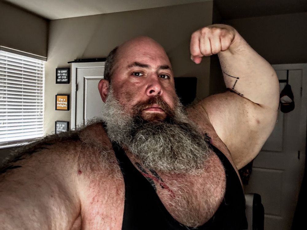 huge man with a bold head and a long greying beard in a tanktop thats taking a selfie with a single bicep pose. It has been morphed to be a realistic 20% larger than original. 