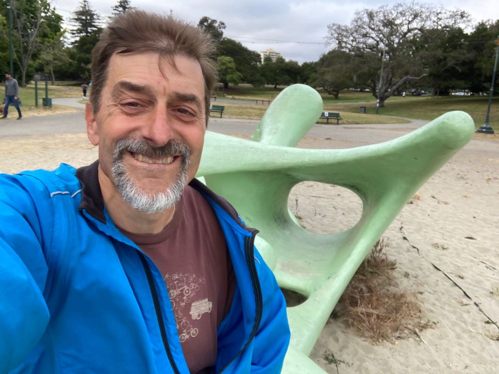 A selfie of a smiling man in front of an abstract modernist sculpture, curves and swoops painted in green. Students of early funk will recognize it as the sculpture the band posed on on the cover of Sly and the Family Stone’s “Dance to the Music”
