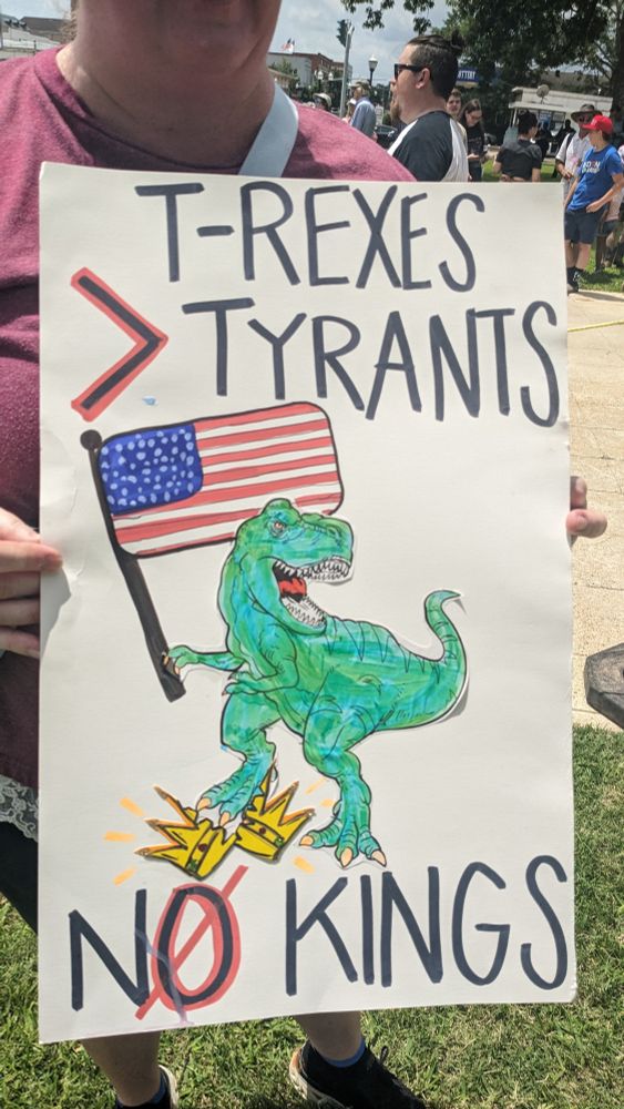A poster with a picture of a T-Rex cracking a crown in two while holding an American flag. The text around it reads T-Rexes > Tyrants, NO KINGS.