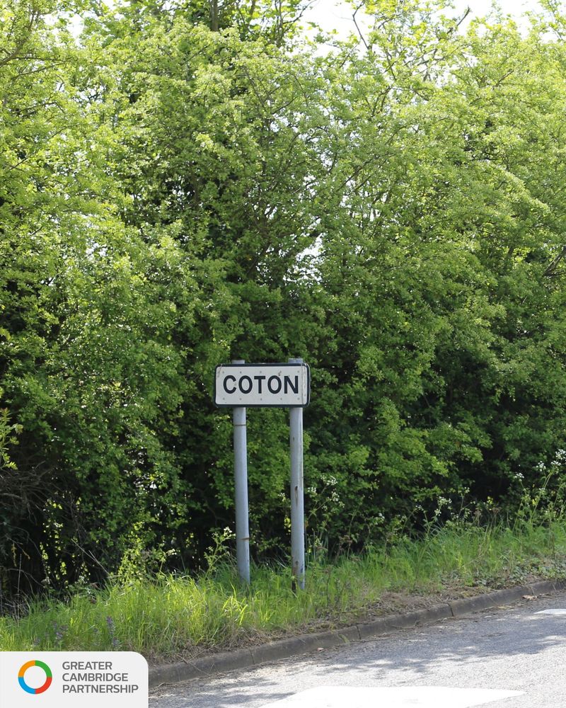 Roadside village sign reading 'Coton' in black letters on a white background, mounted on two grey posts, with dense green foliage and trees in the background. The Greater Cambridge Partnership logo is visible in the bottom-left corner.