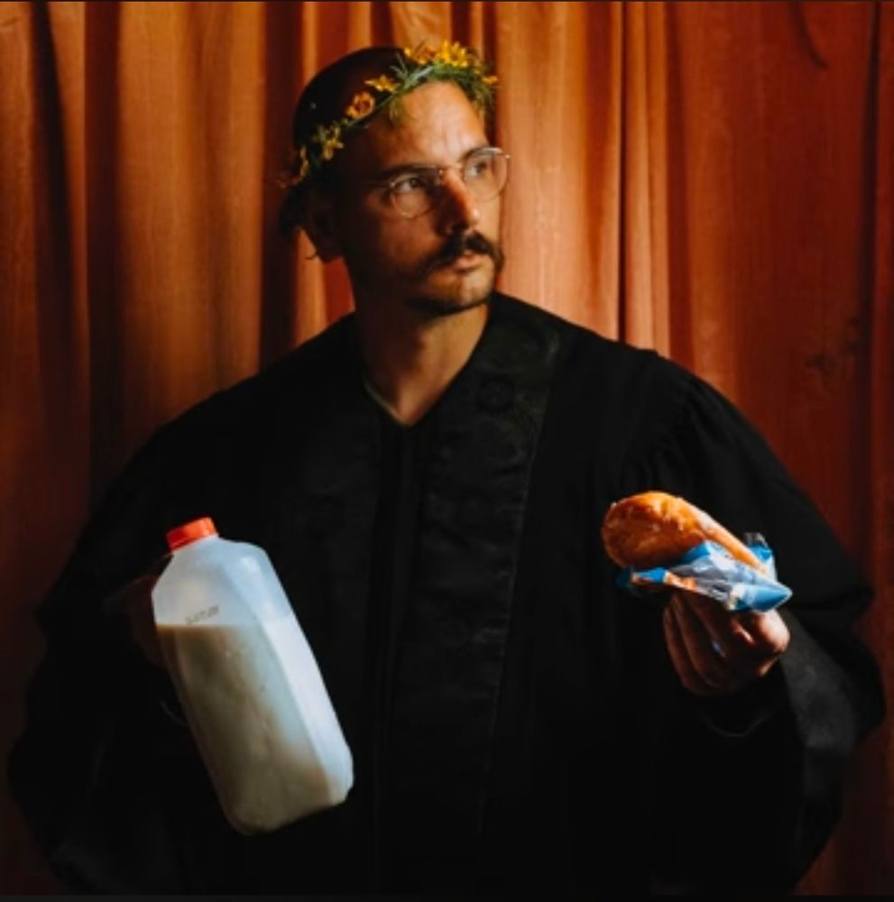 In front of a faded, burnt orange, curtain backdrop, Country Folk Artist Nathan Evans Fox is wearing a flower crown and black judge's robes while holding a partially drunken half-gallon jug of whole milk in his right hand and an open honey bun in his right.