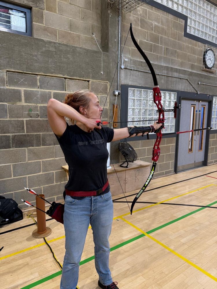 The author doing archery. The form isn’t perfect, but she’s back after time off. The bow is red and black, as is her quiver. Her tshirt says bog witch.
