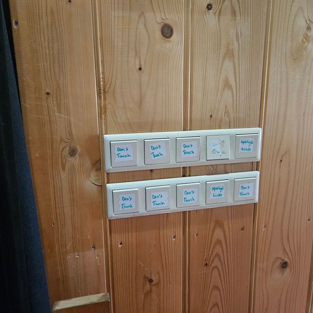 A photo of 10 light switches on a wooden wall.   Eight of them have a sticker pasted on them that reads "don't touch". One reads "spotlight links" - links is Dutch for left, and one "spotlight rechts" - rechts is Dutch for right.