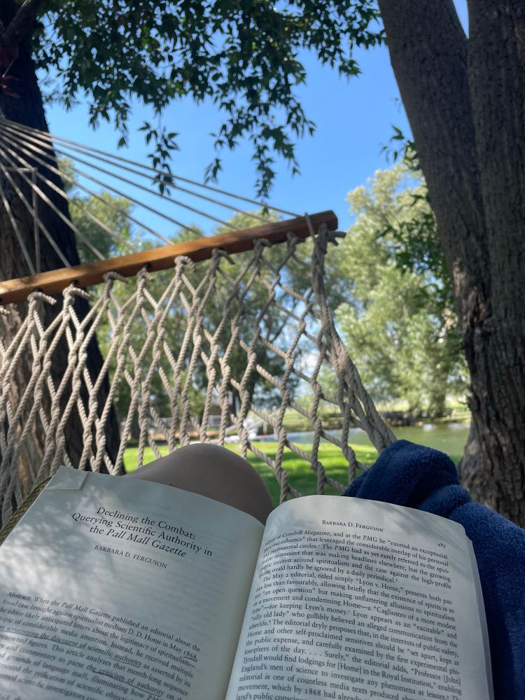 Photo of a hammock near a pond. A person’s knee is visible behind an open academic journal. 