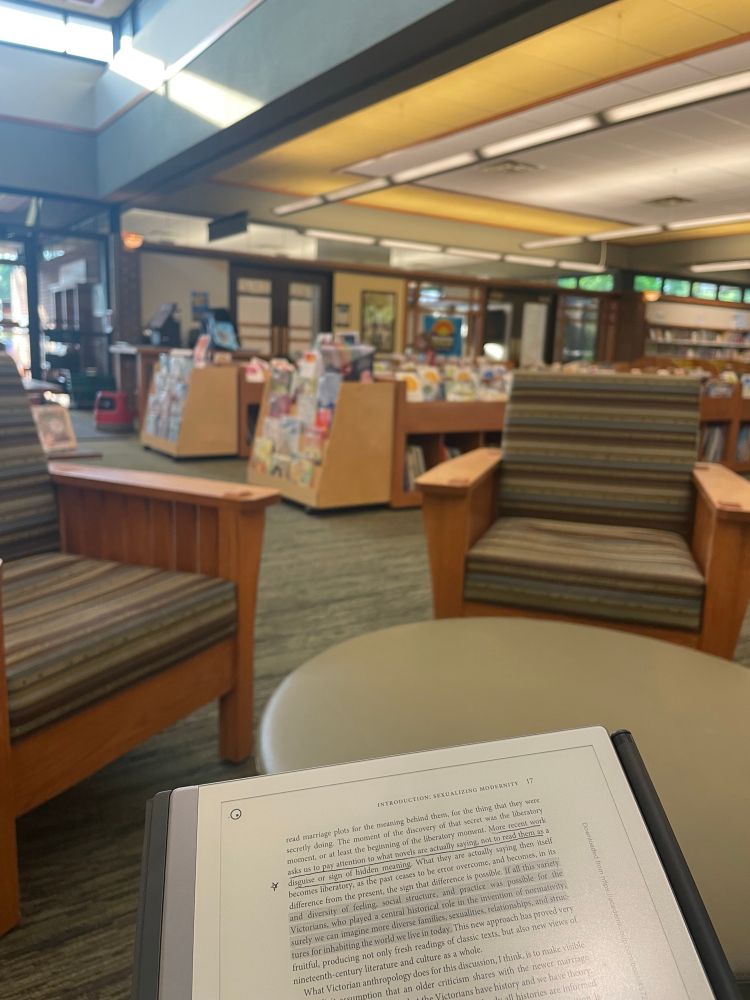 A digital tablet shows annotated text in the foreground. The mid- and background are blurry and show low library shelves and glass doors to a conference room. 