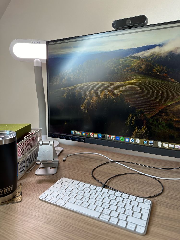 Стіл. На передньому плані лежить біла клавіатура, підключена дротом для зарядки, ближче до стіни висить монітор з веб-камерою на горі, за монітором ховається біла лампа з розсіювачем