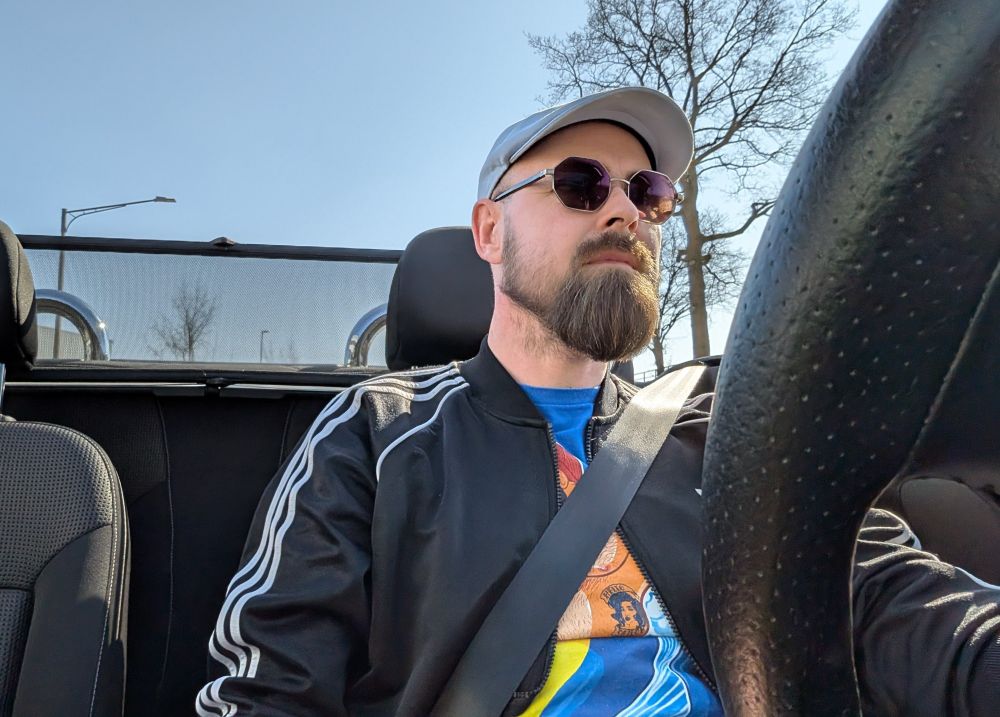 End-40er mit Bart, Sonnenbrille, hellblauem Flatcap, schwarzer Trainingsjacke sitzt am Steuer eines Cabrios. 
