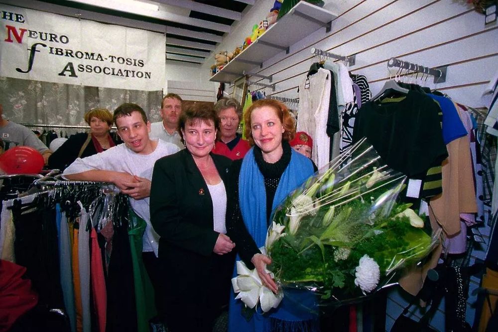 Gillian Anderson poses for a photo with a group of people in a charity shop. She is holding a very large bunch of flowers. 