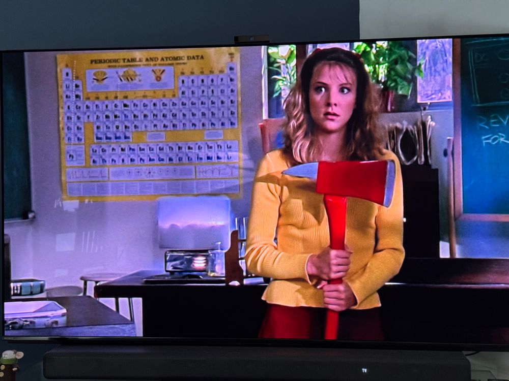 Photo of a tv. Onscreen a woman holds an axe with a periodic table visible behind. 