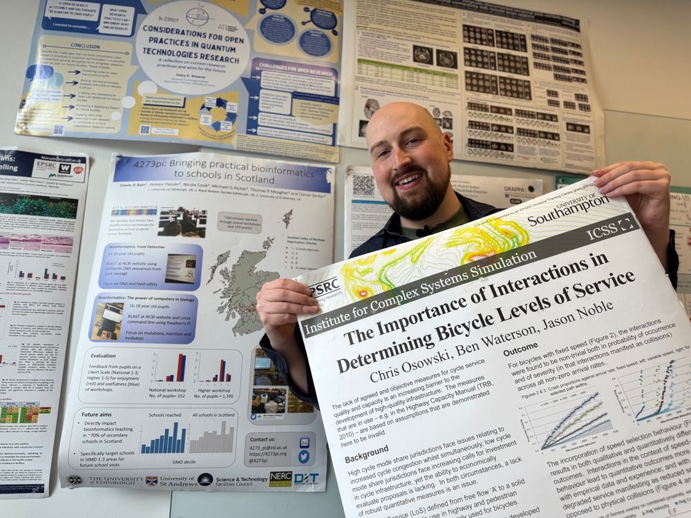 Jamie standing in front of many academic posters. He is also holding one. 