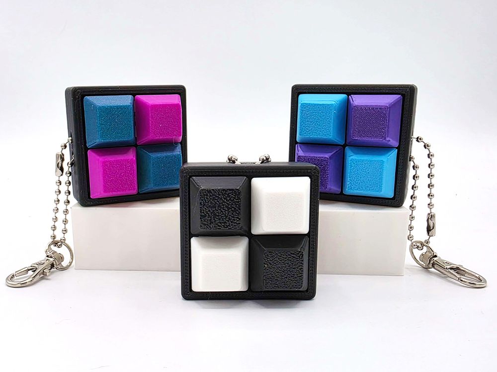 Four square keychains. Inside each are 4 clicky keyboard keys, set in a checkerboard pattern with two of each color: pink and teal, blue and purple, and black & white.
