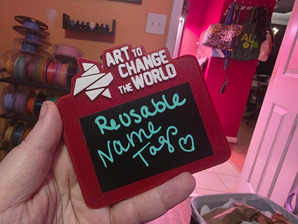 A red rectangle with a white logo at the top that reads Art to Change the World. Underneath is a 2" x 3" black chalkboard sticker. I've written "reusable name tags" on it in green chalk marker.