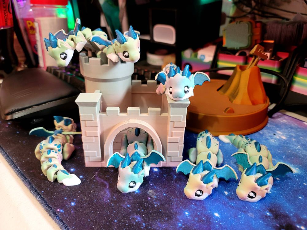 A small cartoonish 3d printed castle with an open front gate and a little tower. 7 tiny chibi-style dragons printed in light pastel green/pink with teal wings and horns ane hanging around the castle or on the castle walls or coming out of the towers.
