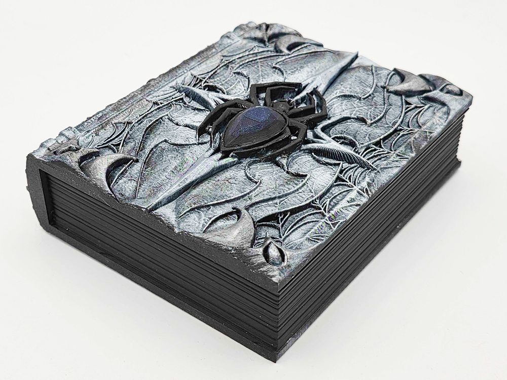 A box that is shaped like a book with a spider motif on it. The webs and box itself are brushed with silver, and the spider is a glossy black with a sparkly gem body. The pages on the book are black, as befits a gothy queen.
