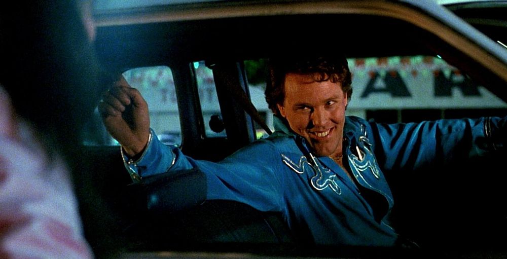 A curly haired man in a blue cowboy shirt, grinning behind the wheel of a car