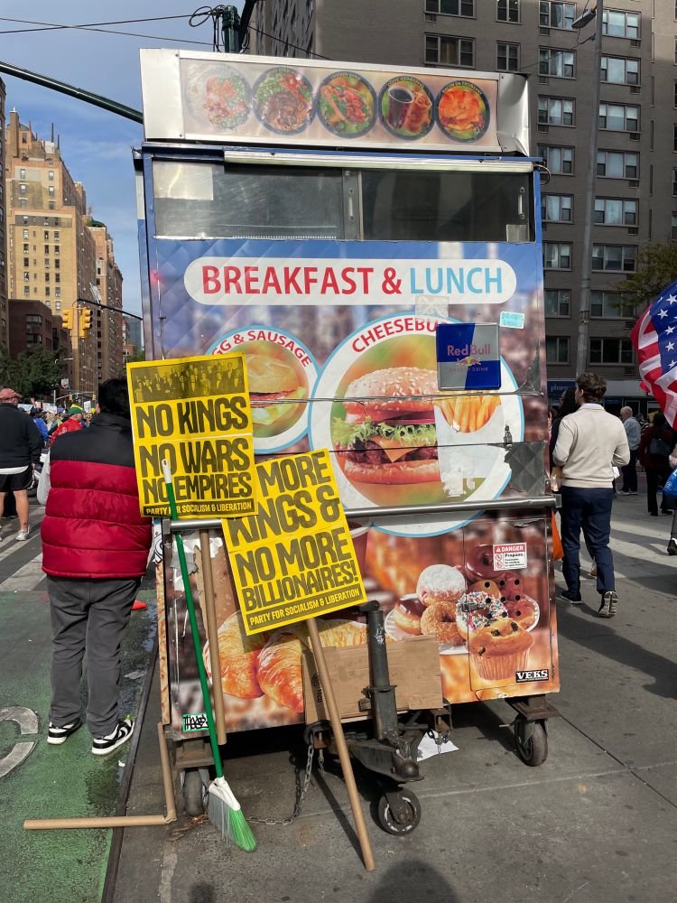 Yellow “no kings” signs leaning against a metal food cart on the sidewalk 