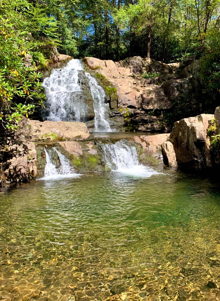 A cascade of waterfalls goes down the cliffs, surrounded by woods on a sunny day. The pool below the falls glows bright light brown-green, the water crystal clear and the rocky bottom visible.